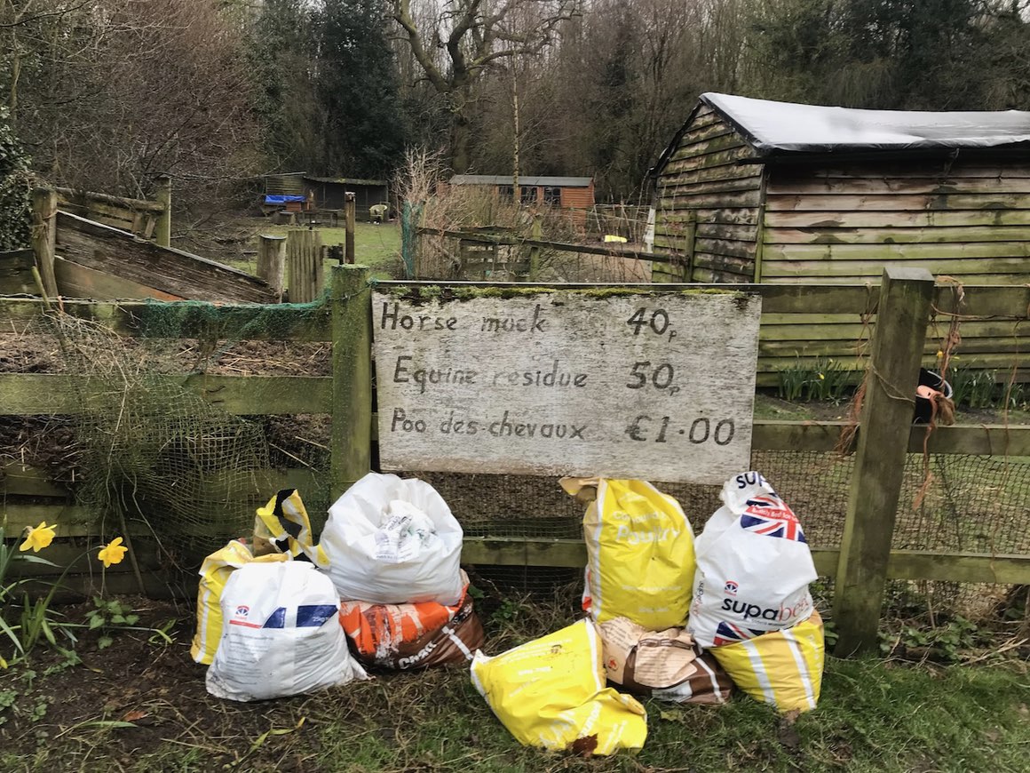 photo of a rural smallholding with wooden sheds, fences, plastic bags of manure, and a handwritten sign that reads: horse muck 40p, equine residue 50p, poo des chevaux £1